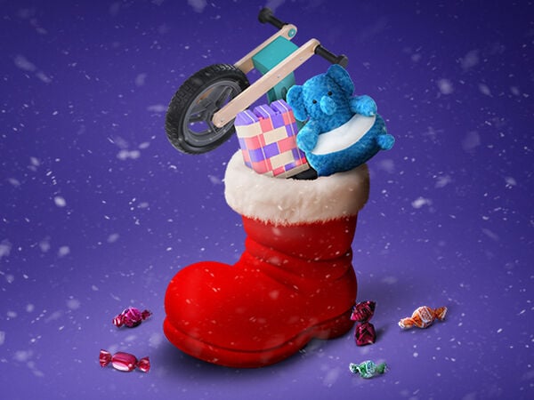 Scenă festivă de Crăciun cu o cizmă mare roșie, cu margine albă de blană, plină cu cadouri, pe un fundal mov acoperit de zăpadă. În interiorul cizmei se află o mică bicicletă de echilibru din lemn, un elefant de pluș albastru și un set colorat de cuburi de construcție pentru copii. În jurul cizmei, pe zăpadă, sunt împrăștiate bomboane colorate ambalate. Fulgi de zăpadă cad în aer.