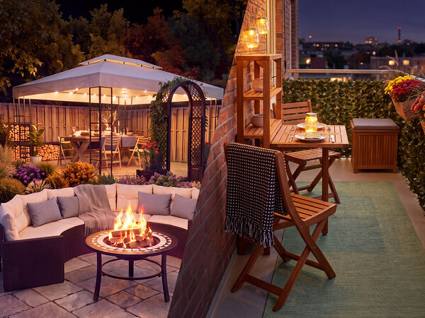 Spații de relaxare &icirc;n aer liber la amurg, cu un set lounge de grădină cu vatră de foc și, pe o parte, un pavilion plin cu flori, iar pe cealaltă parte un balcon decorat cu mobilier din lemn, lumini de la felinare și flori &icirc;n ghiveci, cu vedere spre orizontul orașului.