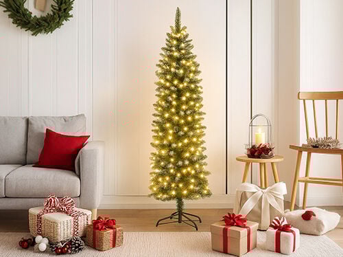 O scenă luminoasă și modernă de sărbători, cu un brad de Crăciun înalt și suplu, preiluminat cu lumini calde albe, amplasat în centru. În stânga se văd brațul și spătarul unei canapele gri deschis, cu o pernă roșie. O colecție de cadouri împachetate, legate cu panglici roșii și albe și decorate cu conuri de pin, se află pe un covor deschis la culoare în prim-plan. În dreapta, un scaun din lemn și o măsuță rotundă mică din lemn susțin o lanternă cu o lumânare, completând atmosfera caldă și festivă.