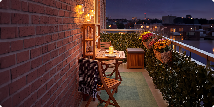 Balcon mic cu scaune și masă din lemn, luminat de felinare și lumânări, decorat cu flori în ghiveci și verdeață, cu vedere spre orizontul orașului pe timp de noapte.